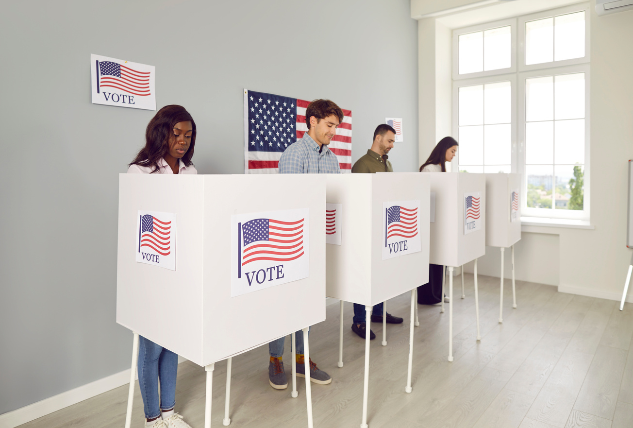 American citizens standing at vote center in voting booth putting ballots in bin on election day.